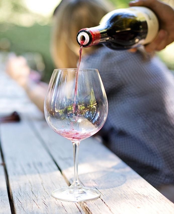 A hand pouring red wine from a bottle into a clear wine glass placed on a rustic wooden table outdoors, with blurred people in the background.