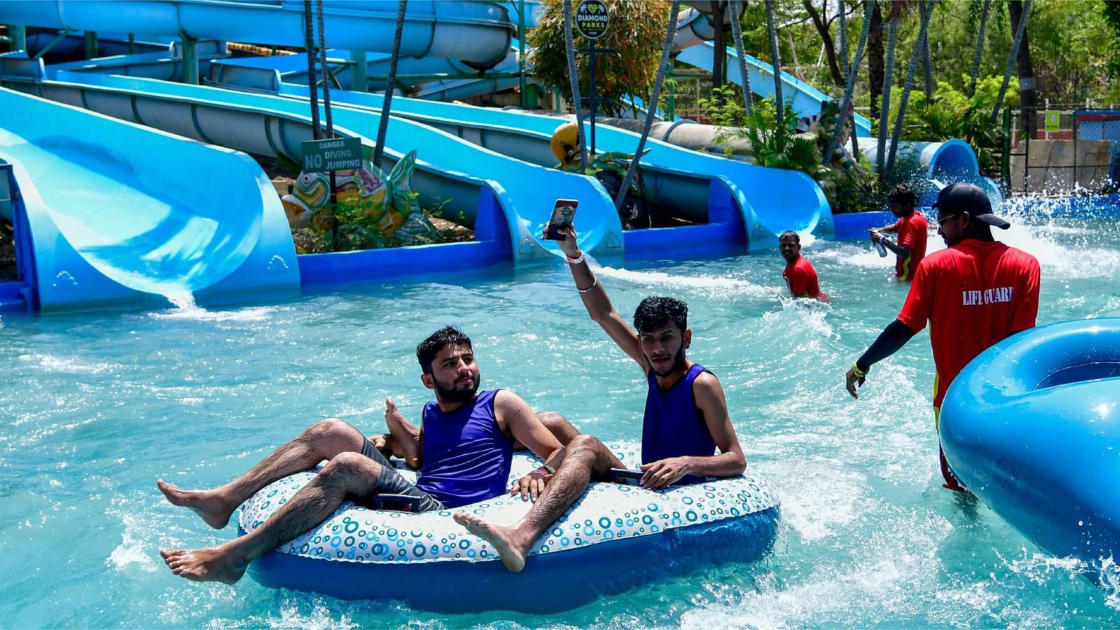 Two people riding inflatable tube through the wave pool at Diamond Parks, Pune, with splashing water and blue water slides visible in the background.