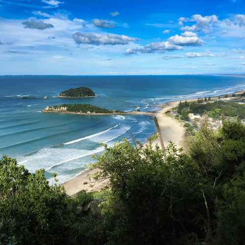Coastal beach with turquoise waves, lush greenery, and a small island under a partly cloudy sky.