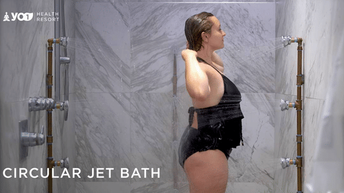 A woman in a dark swimsuit stands in a circular jet bath, adjusting her hair at YO1 Longevity & Health Resorts