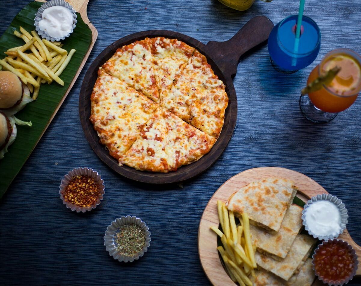 Top view of a table with various fast foods like pizza, fries, burgers, and dips, along with two drinks placed at Oriental Aster, Mumbai.
