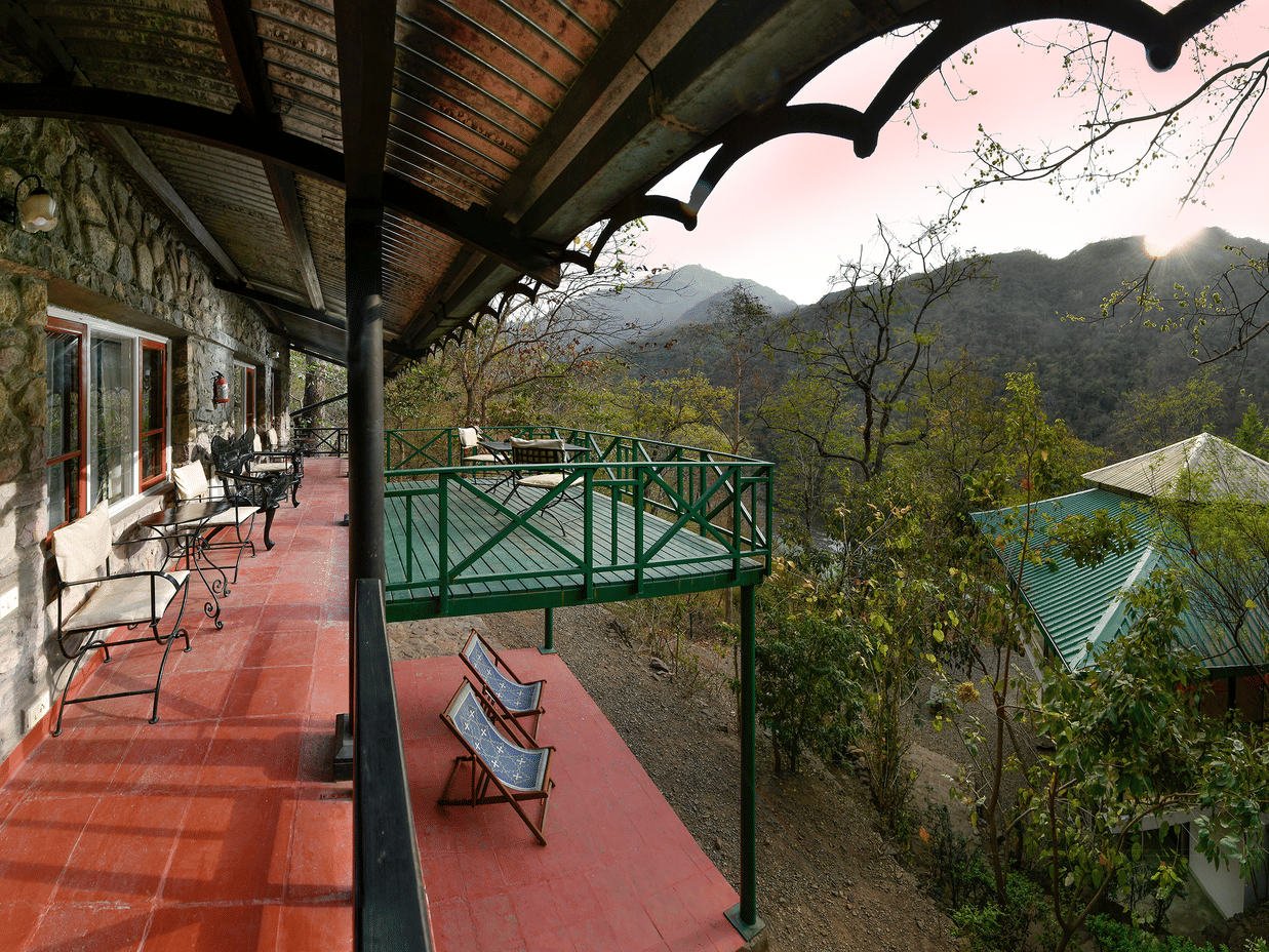 Wide-angle view of a long veranda with a red floor and green wooden railings looking out over a lush green valley and mountains at Neemrana’s Glasshouse on The Ganges offering a peaceful outdoor space for guests to relax and soak in the scenery.