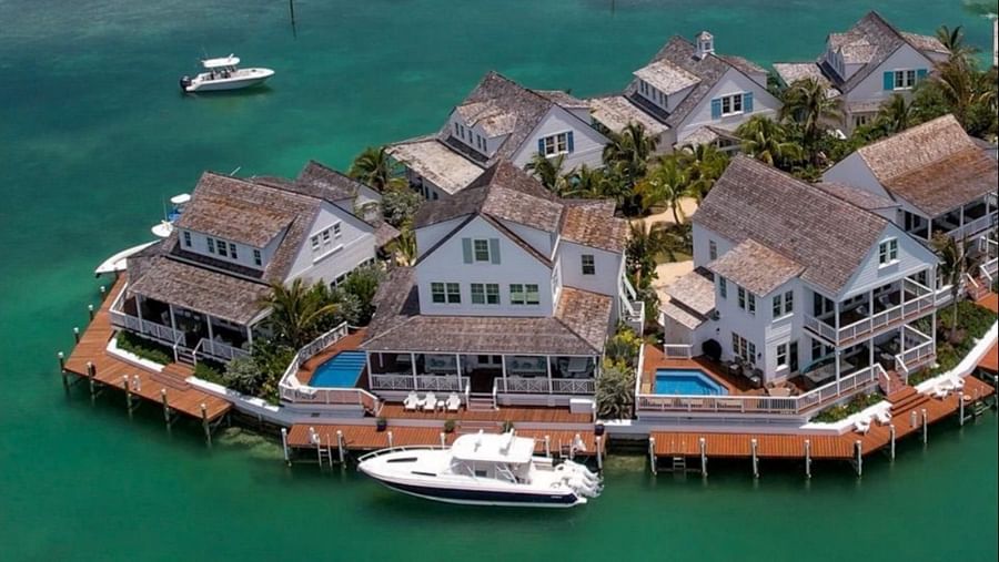 Multiple white waterfront homes with docks, some with pools, on a lush green island in turquoise water, with boats moored and another cruising near Abaco Inn.