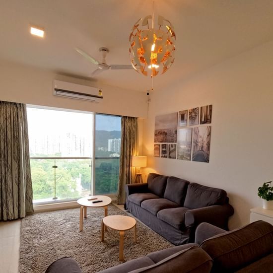 A full view of a living room with a sofa and glass windows at Sahil Exotica by Maxxvalue Apartemnt