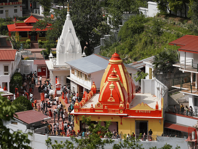 High-angle view of the vibrant red and yellow Kainchi Dham surrounded by white buildings and lush green trees.
