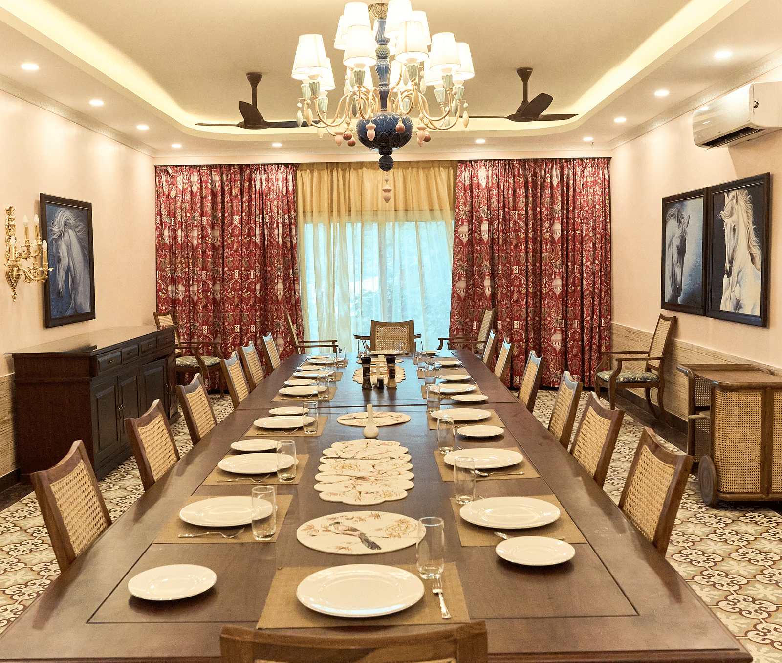 At Mayfair Manor, Alibag, a spacious dining table is beautifully arranged for a delightful culinary experience, enhanced by wall-mounted paintings, an elegant chandelier above, and expansive windows with curtains tastefully drawn to the side
