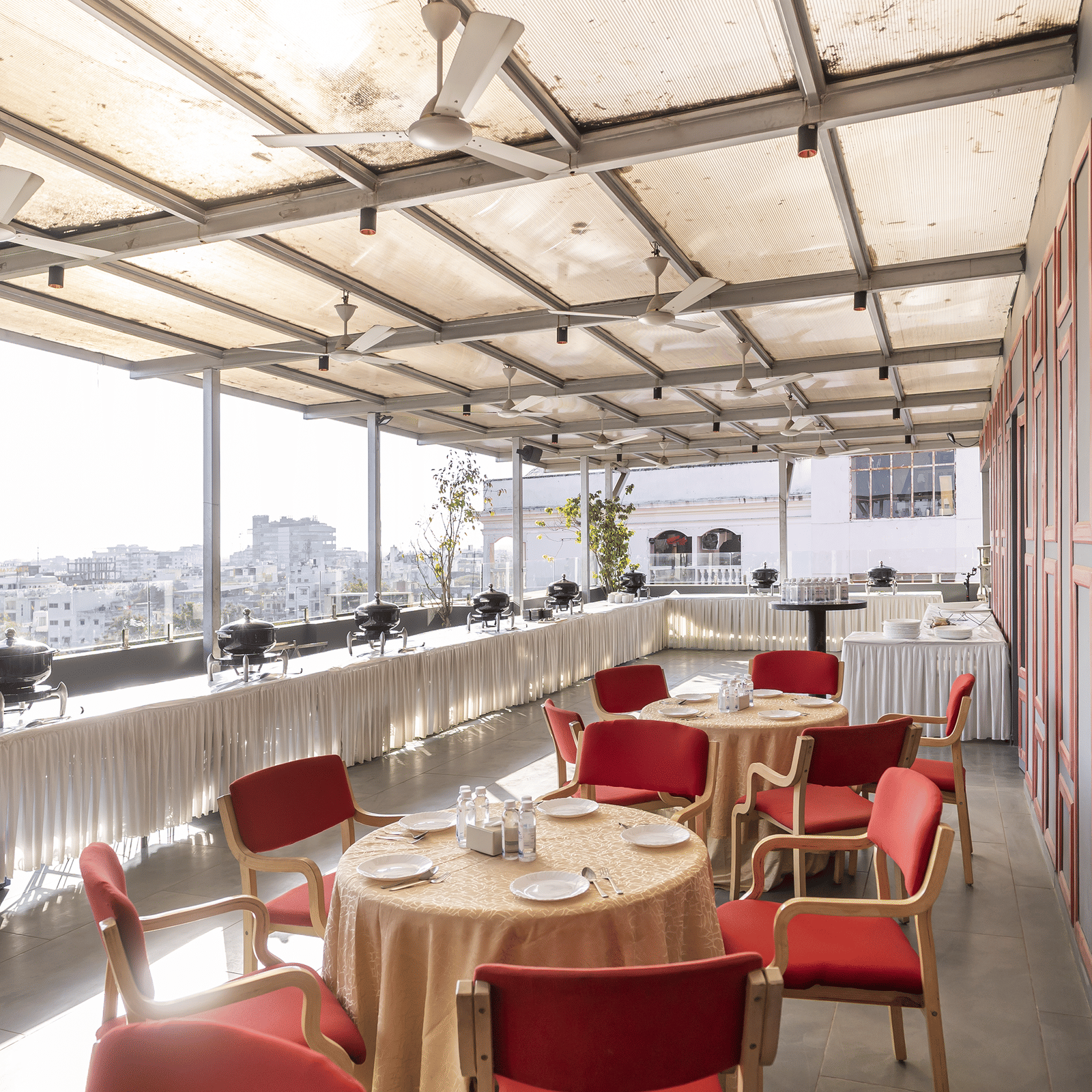An outdoor seating area with round tables, red chairs and a roof covering with a city view at Infiniti Hotel, Indore.