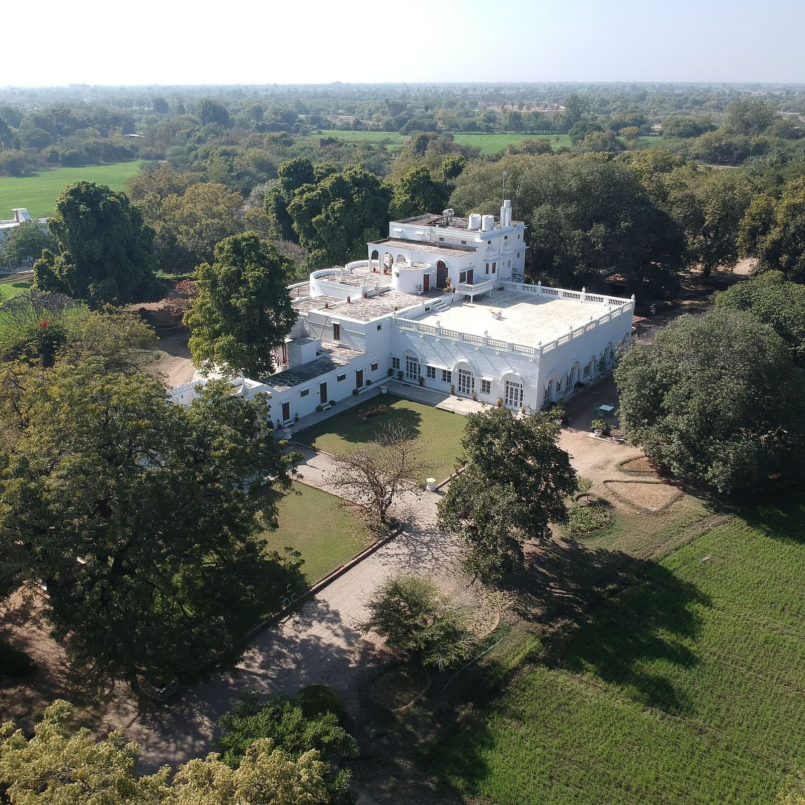 An aerial view of a white building complex with a central courtyard, surrounded by lush greenery and fields- Shahpura Bagh.