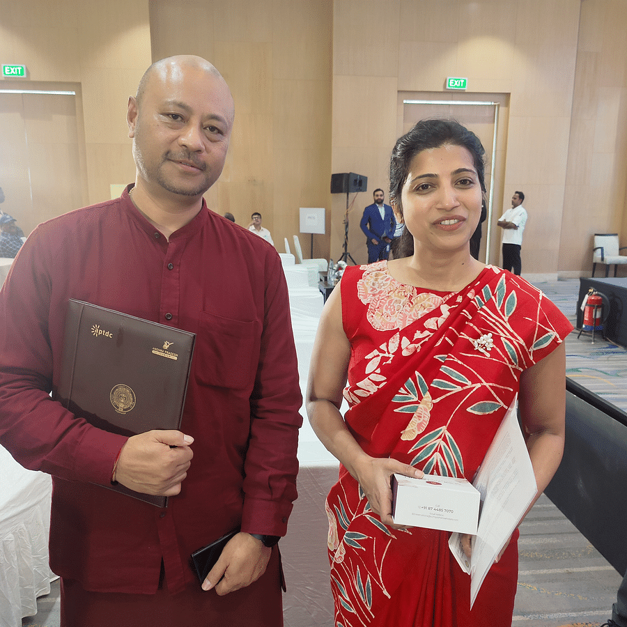A man in a maroon outfit holding a signed document and a woman in a red saree holding papers and a box, both smiling at the camera at a formal event.