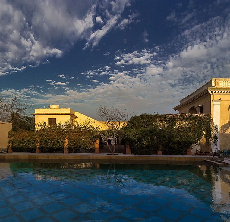 A wide view of the Royal Heritage Haveli grounds with a pool reflecting the sky and architectural elements under a cloudy sky.