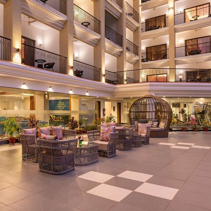A spacious, well-lit hotel atrium with multiple floors of balcony rooms overlooking a central seating area featuring stylish wicker egg chairs, round tables, and decorative plants - Golden Tulip Candolim.