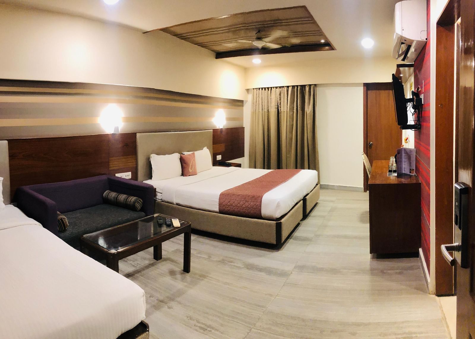 Hotel room interior showcasing a double bed, a dark-colored sofa, a glass-topped coffee table, and a work desk with a chair  | Astra Hotels and Suites Kadubeesanahalli
