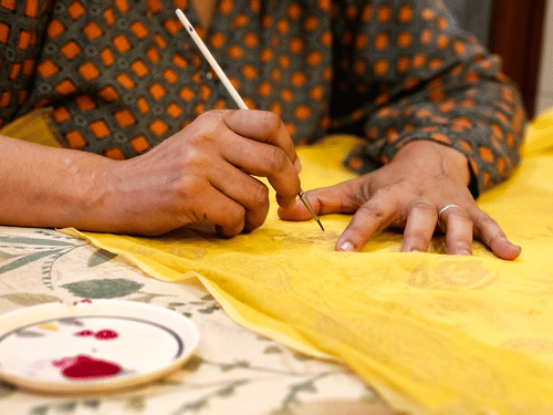 An artisan uses a fine brush to carefully paint intricate designs onto bright yellow cloth.
