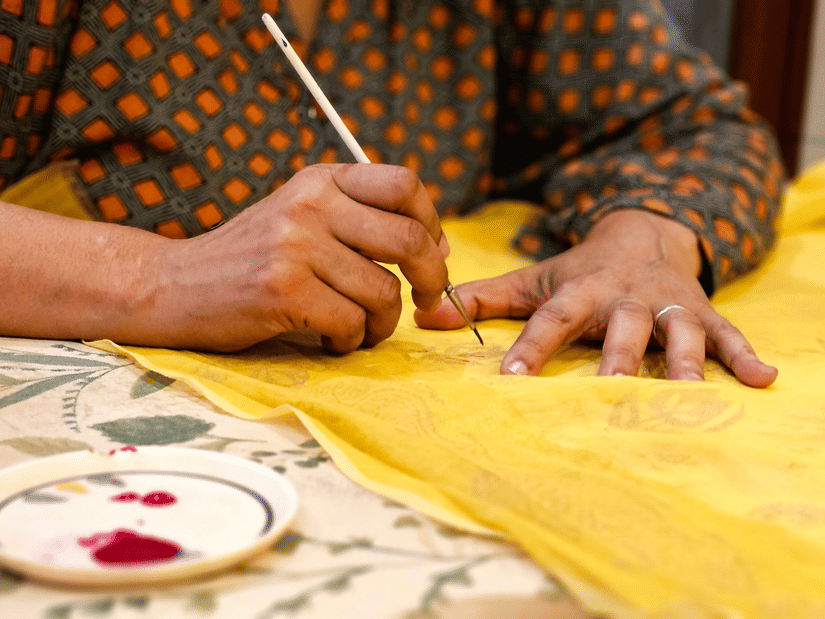 An artisan uses a fine brush to carefully paint intricate designs onto bright yellow cloth.