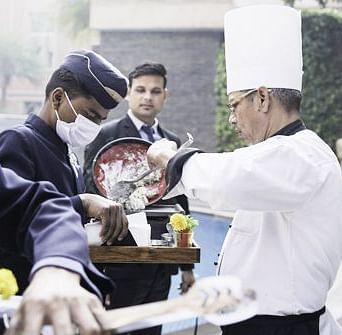 A chef working on a dish held by the staff with a man in the background at Nirvana Luxury Hotel, Ludhiana.