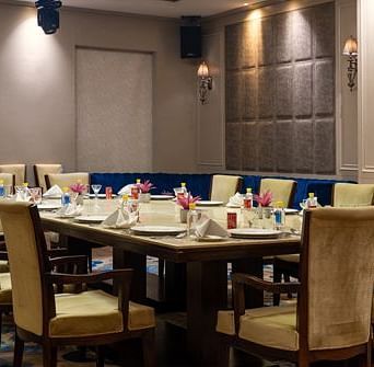 A warmly lit private dining space at Nirvana Luxury Hotel, Ludhiana, showcases a long, set table with elegant chairs and a comfortable blue banquette seating area.