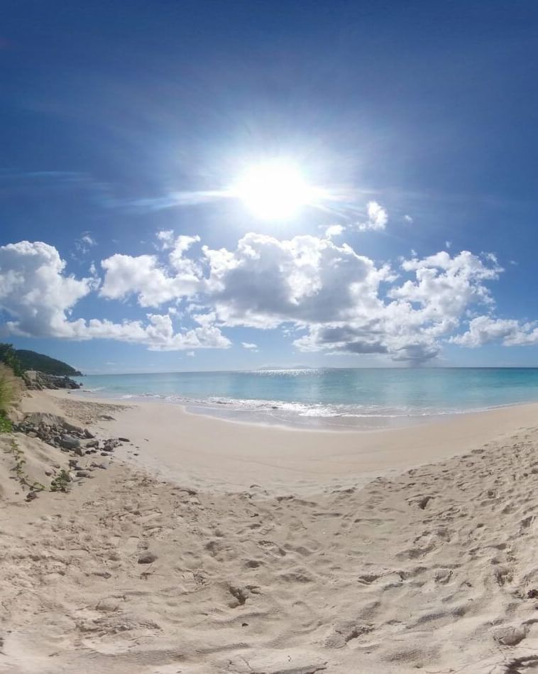 A wide-angle view of a serene, empty white sand beach under a bright blue sky with scattered clouds and sparkling turquoise water.