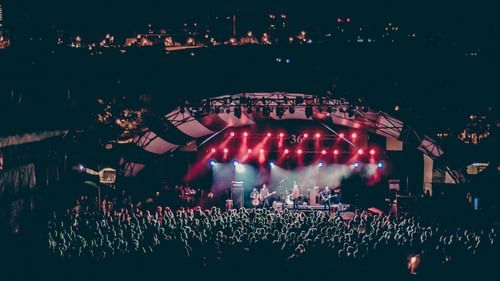 A wide-angle view of a nighttime outdoor concert with a large crowd and a stage lit by vibrant pink and purple lights.