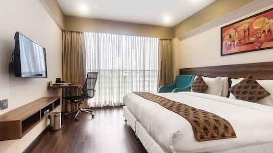 A side view of a bed in a bedroom with a TV on the wall and a chair along with a huge glass window | Mumbai House Hotels