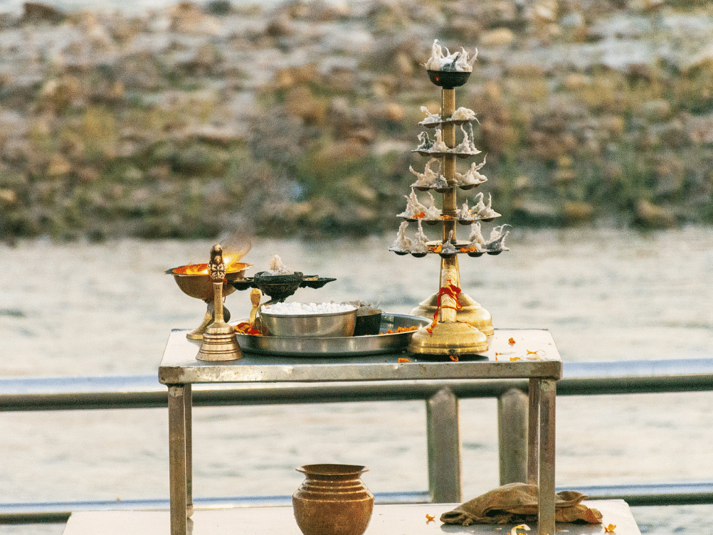 Traditional Indian puja setup on a riverside platform, featuring brass lamps, a kalash, and ritual items.