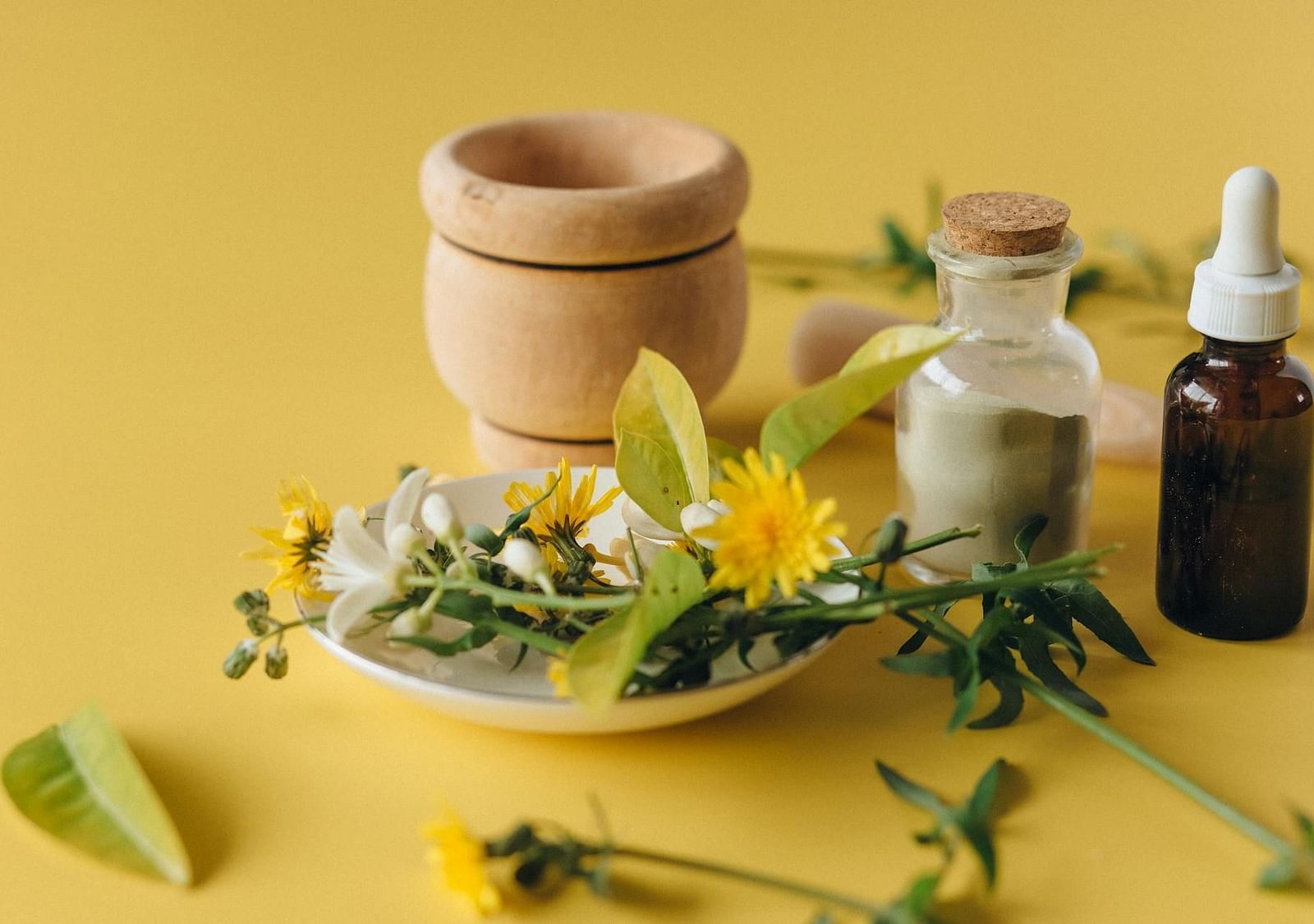 Spa items kept on a surface including flowers, a glass jar with powdered herbs, an essential oil bottle, and a wodden herb crusher