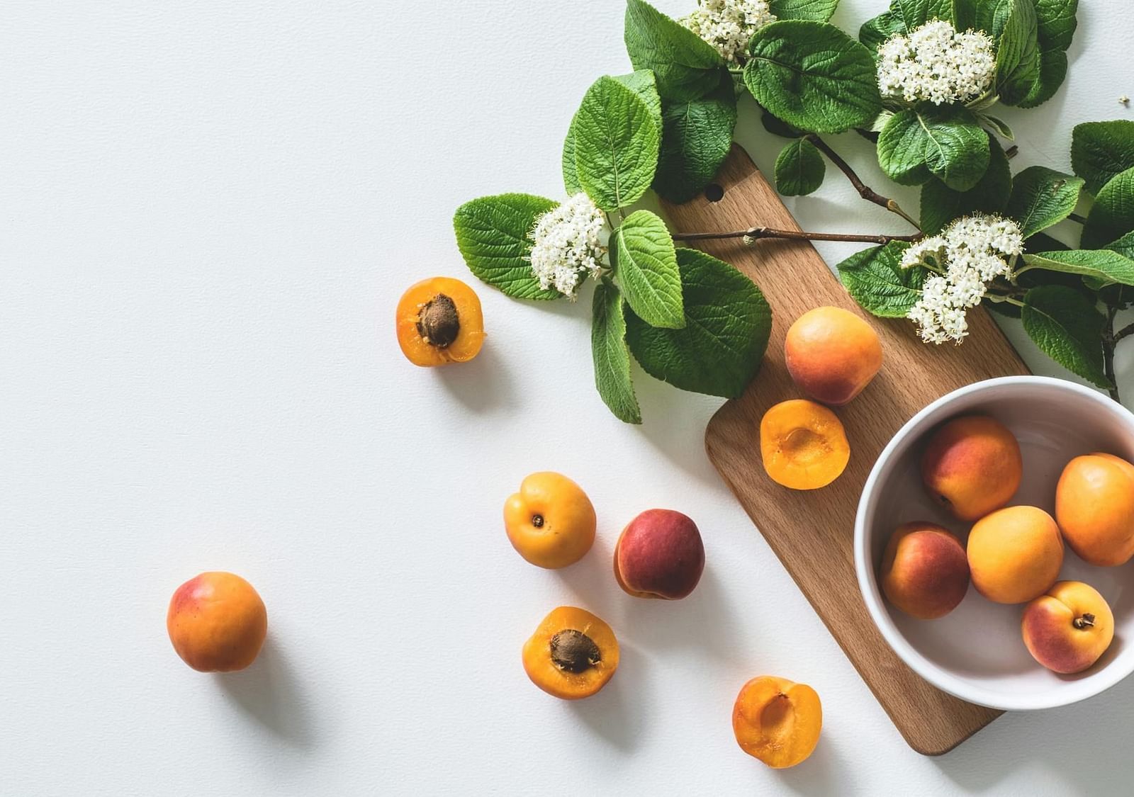 A bowl of peaches with some peaches outside, next to flowers with green leaves