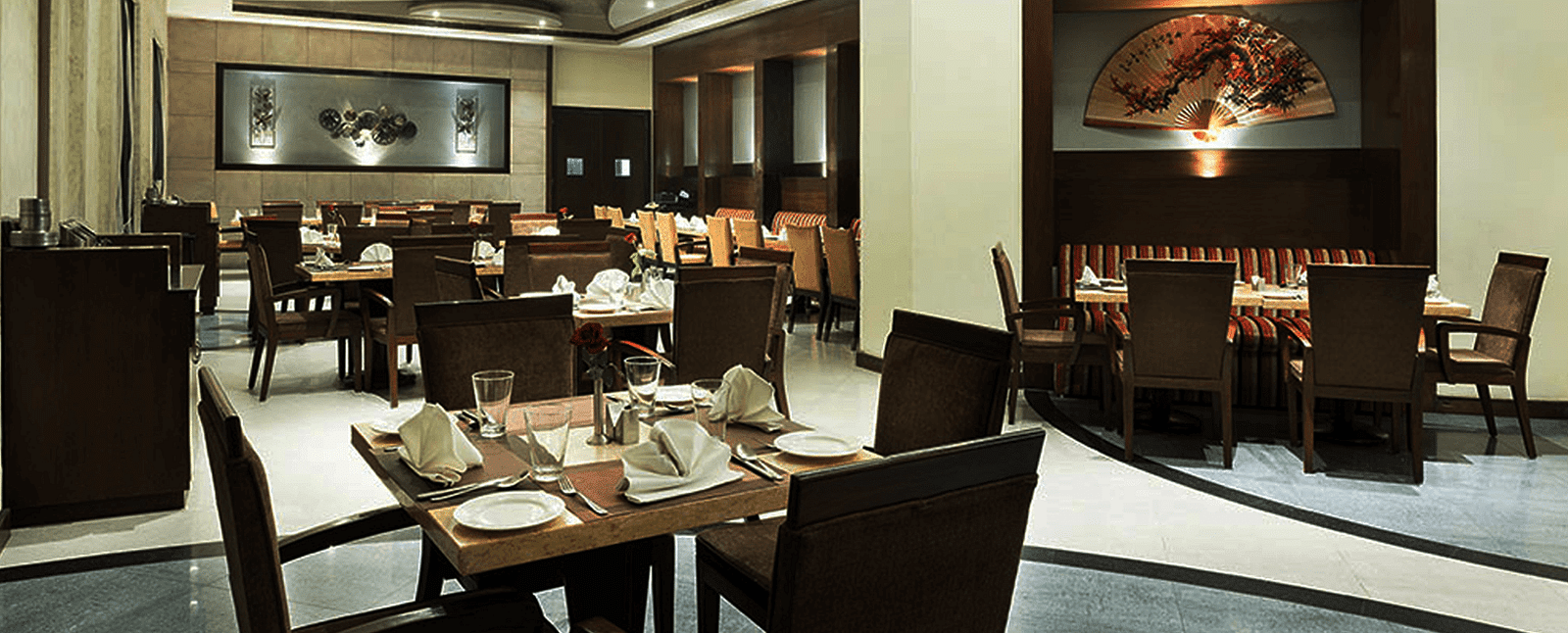The elegant dining area at Nirvana Luxury Hotel, Ludhiana, features neatly set tables with silverware and napkins, surrounded by dark chairs, and accented by subtle lighting and artwork.