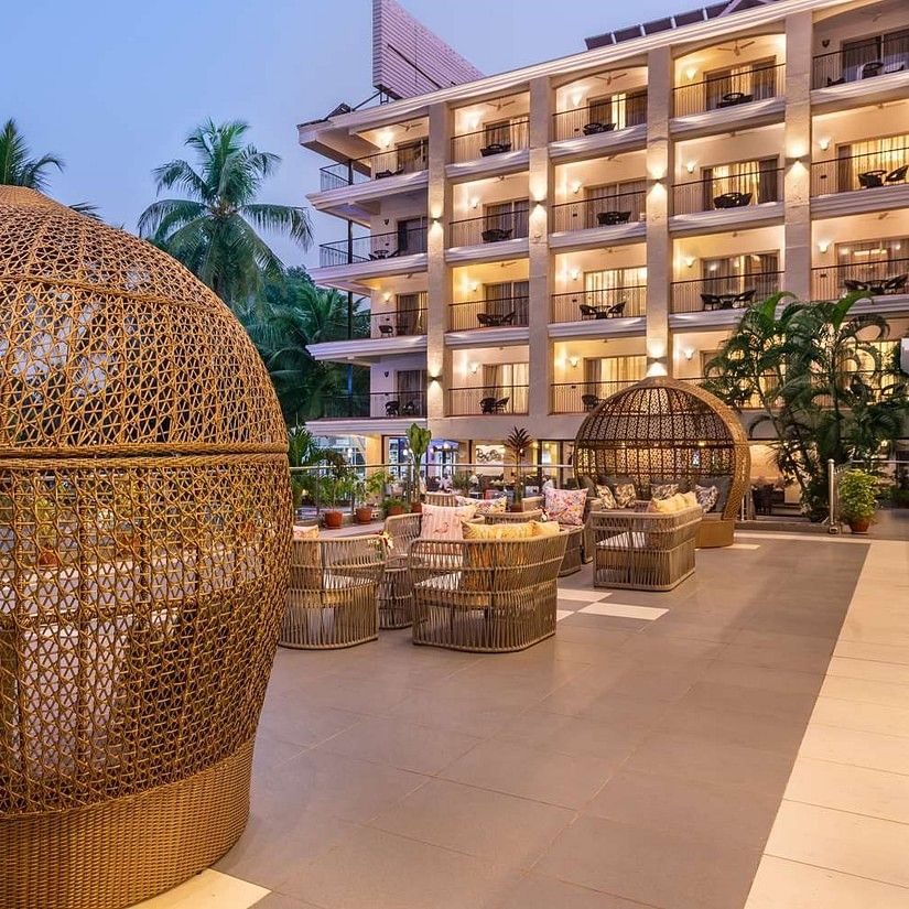 An elegant outdoor lounge area at dusk, featuring large woven egg-shaped seating pods, round tables with chairs, and a multi-story, warmly lit hotel building in the background surrounded by palm trees -  Golden Tulip Candolim.