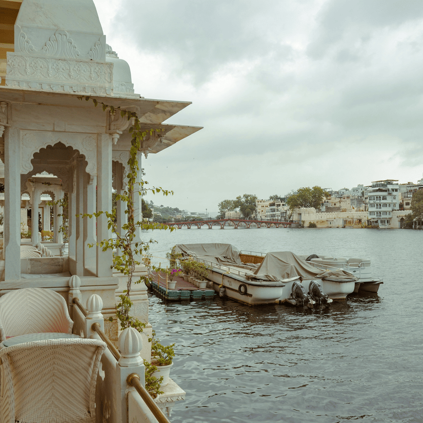 White lakeside corridor with chairs beside calm water and covered boats docked along Lake Pichola