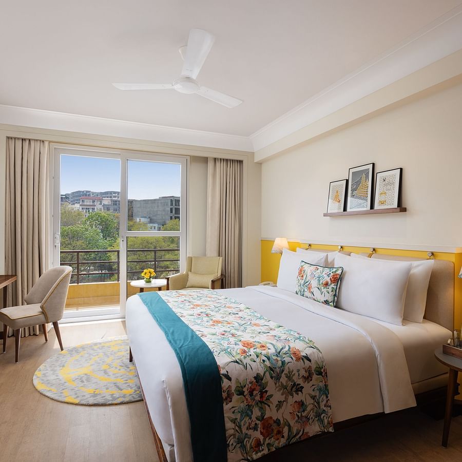 A sunny hotel room with a queen bed dressed in white linens and a floral and teal throw, flanked by a wooden nightstand with a lamp and a small round table, a desk with a chair and television, and a balcony with outdoor seating offering a view of trees and