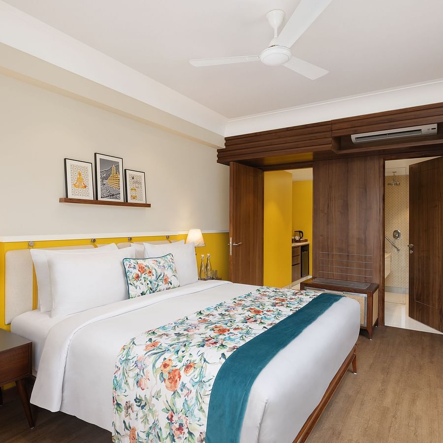 A bright hotel room with a queen bed featuring floral and teal accents, a wooden nightstand with a lamp and phone, a shelf with framed pictures, wooden wardrobes, and an open doorway to a bathroom with yellow walls - Aloha On the Ganges.