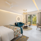 A bright and airy hotel room featuring a queen bed with floral accents, a teal velvet sofa, a round coffee table on a patterned rug, a work desk with a chair and a television, a balcony with outdoor seating and a view of greenery, modern lighting, and a c