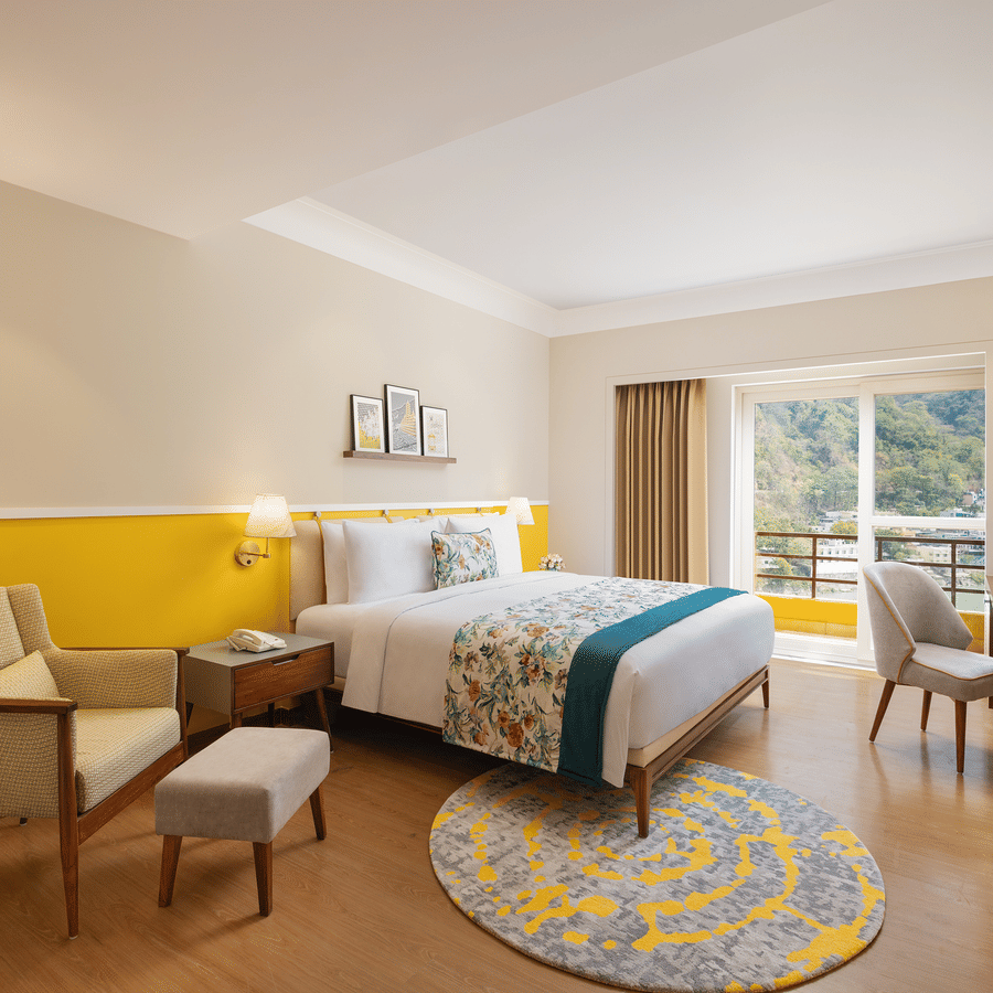 A bright hotel room with a queen bed featuring floral and teal bedding, a comfortable armchair with an ottoman, a wooden desk with a chair and a television, a floor lamp, and a balcony with a scenic view - Aloha On the Ganges.