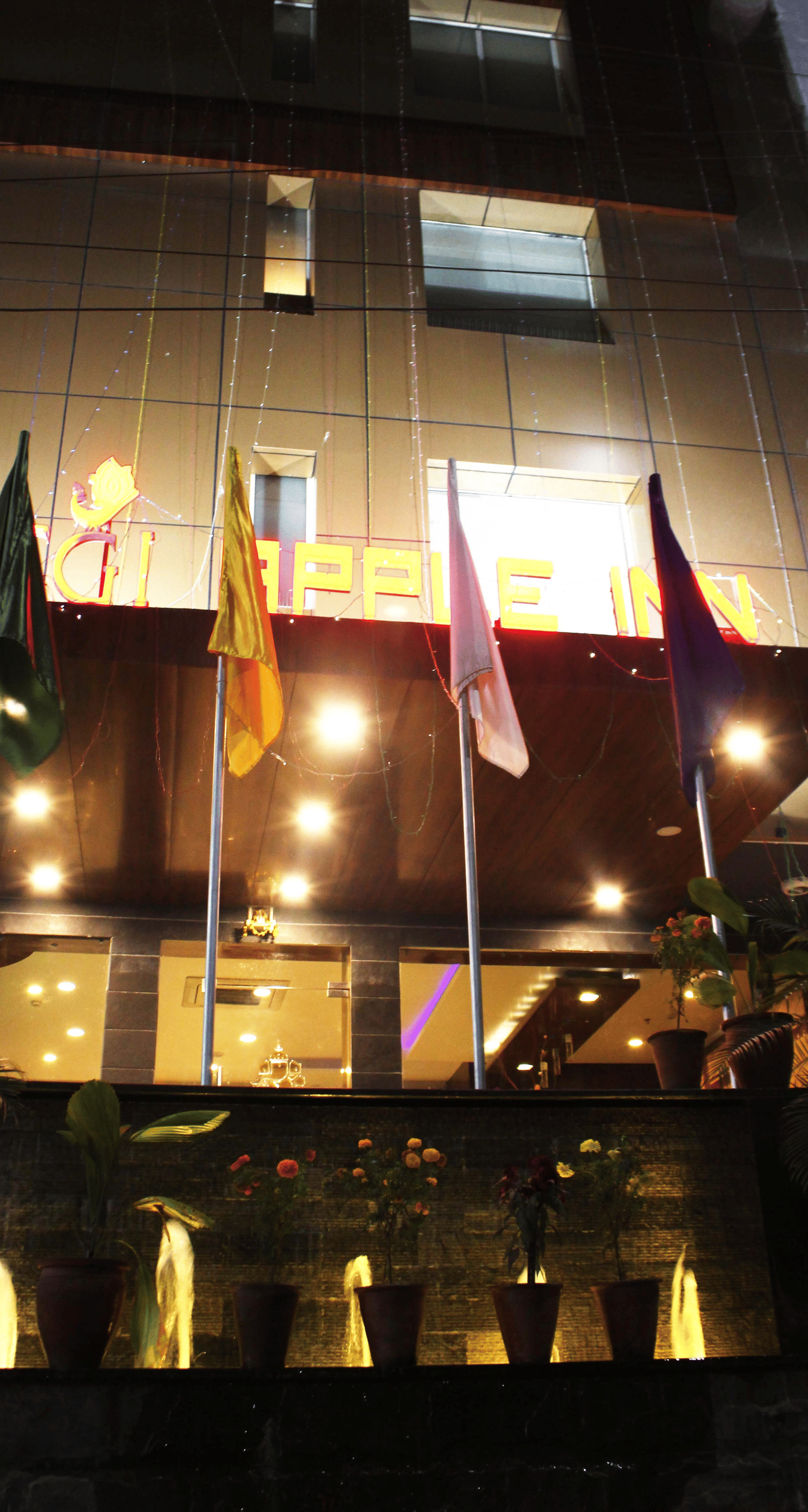 A view of the hotel from the front illuminated by warm yellow lighting and with flags placed in the front - TGI Apple Inn, Jaipur 