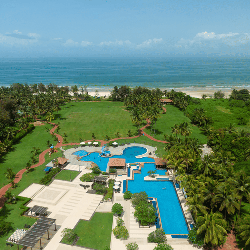An aerial view of Kenilworth Resort & Spa, Goa, with vast blue ocean at the far end and a swimming pool, a lawn area, pathways and many trees surrounding the property. It is the best place for a long stay in Goa.