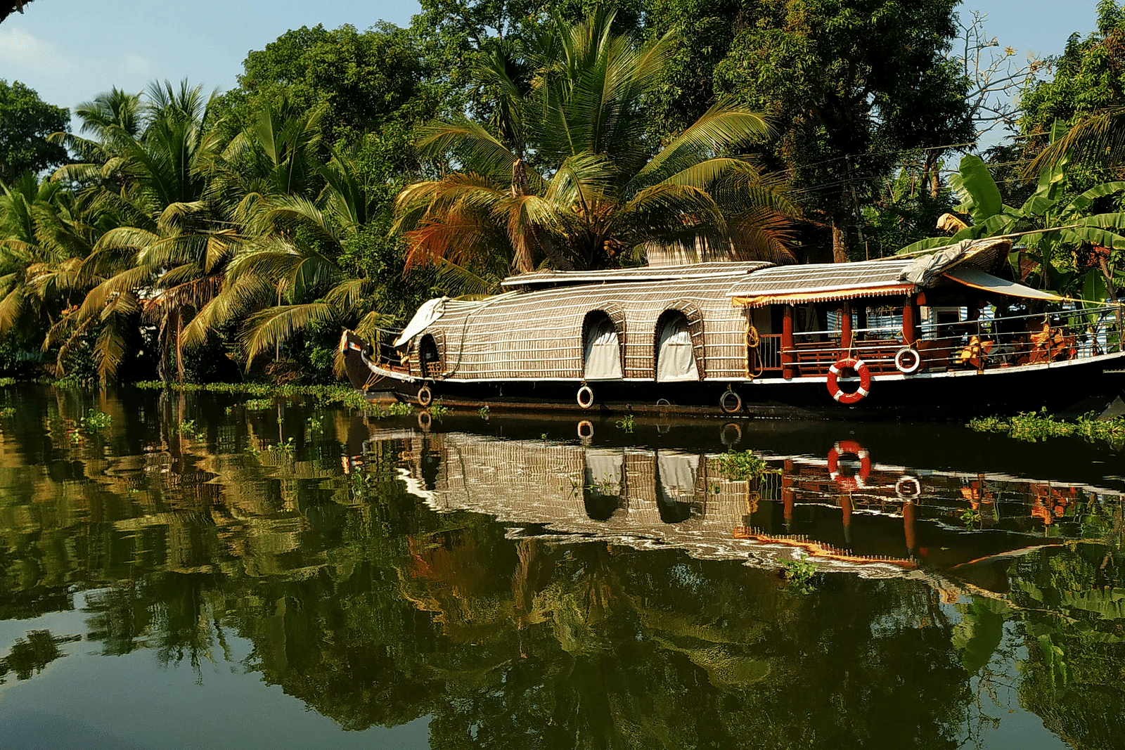 A traditional houseboat with a thatched roof is floating on the calm backwaters, flanked by lush greenery and palm trees reflecting in the water.