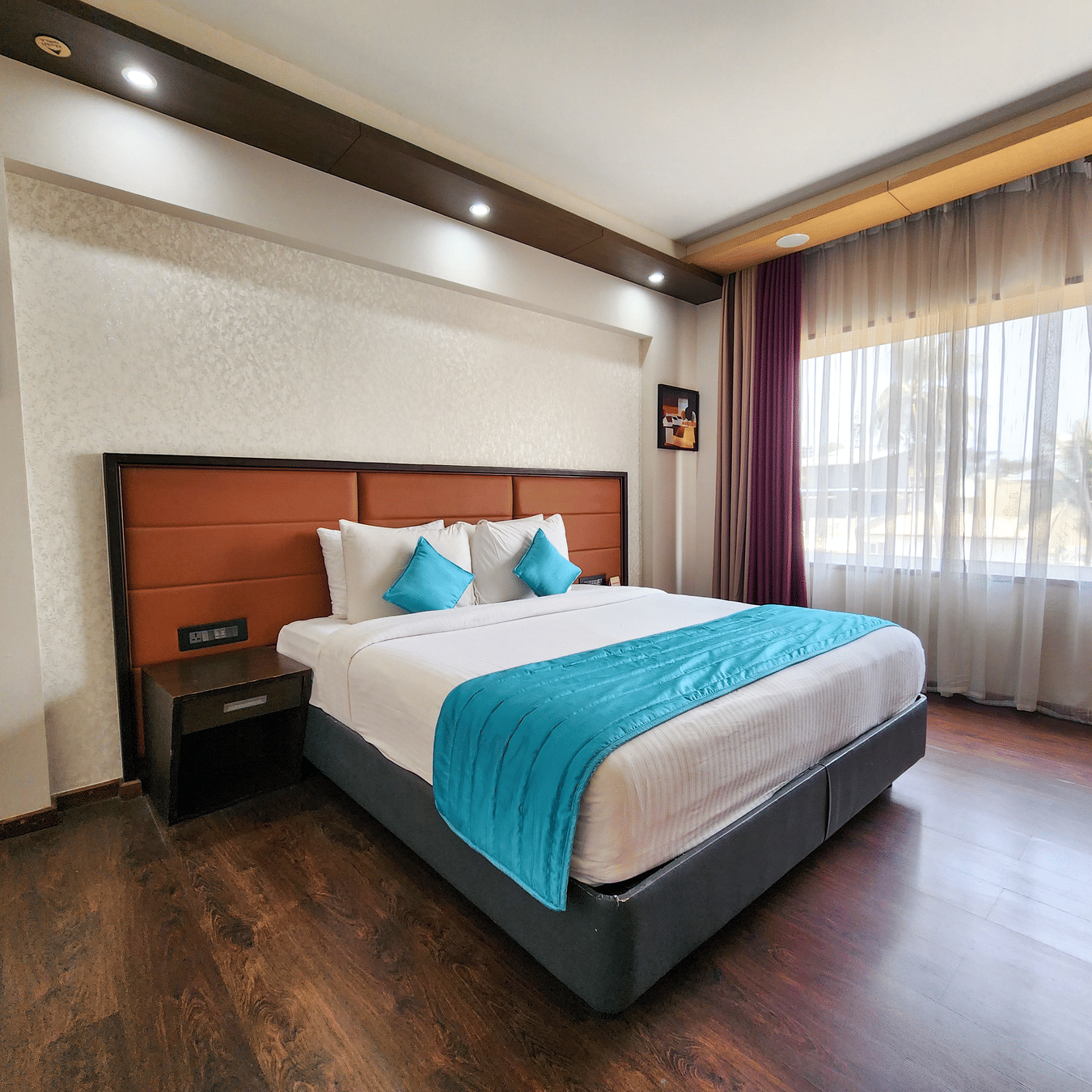 A spacious room at Dimora, Thiruvananthapuram, featuring a well-made bed, a headboard, a bedside table, and daylight streaming in through sheer curtains.