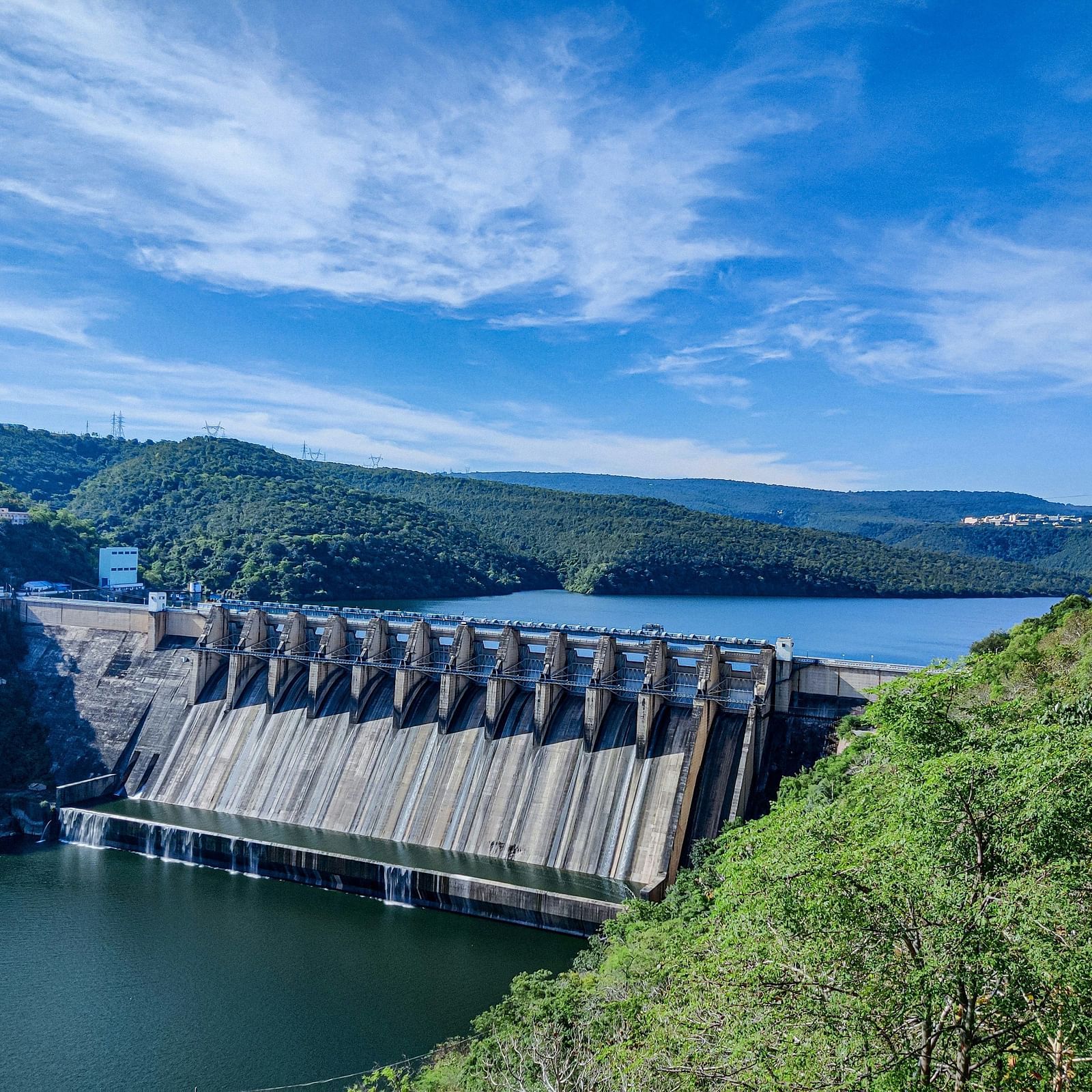A scenic view of a large dam with a reservoir surrounded by lush green hills and a clear blue sky, showcasing a blend of engineering and natural beauty.