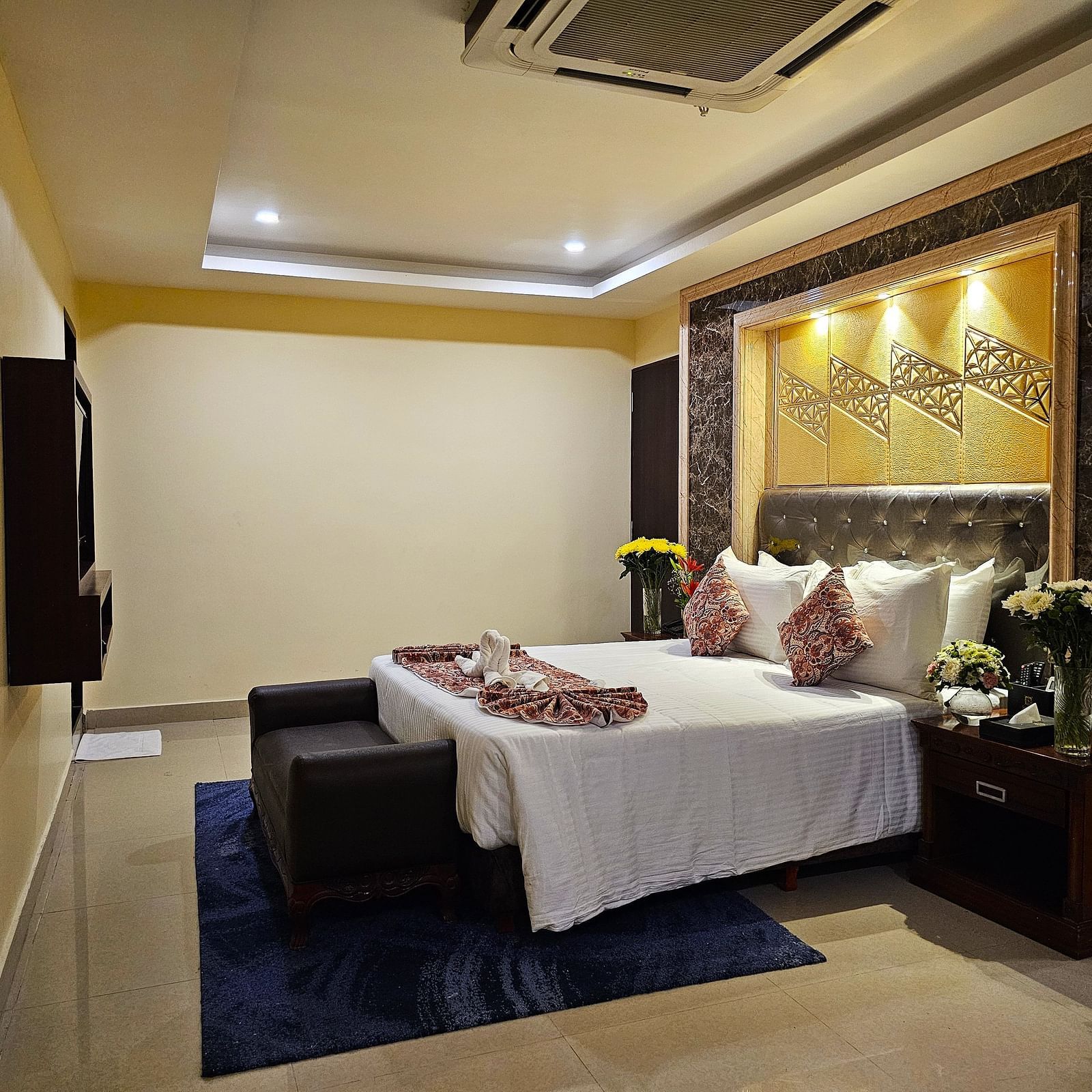 A bedroom featuring a plush bed next to a nightstand facing the TV mounted on the wall under warm lighting at Sathyam Grand Resort