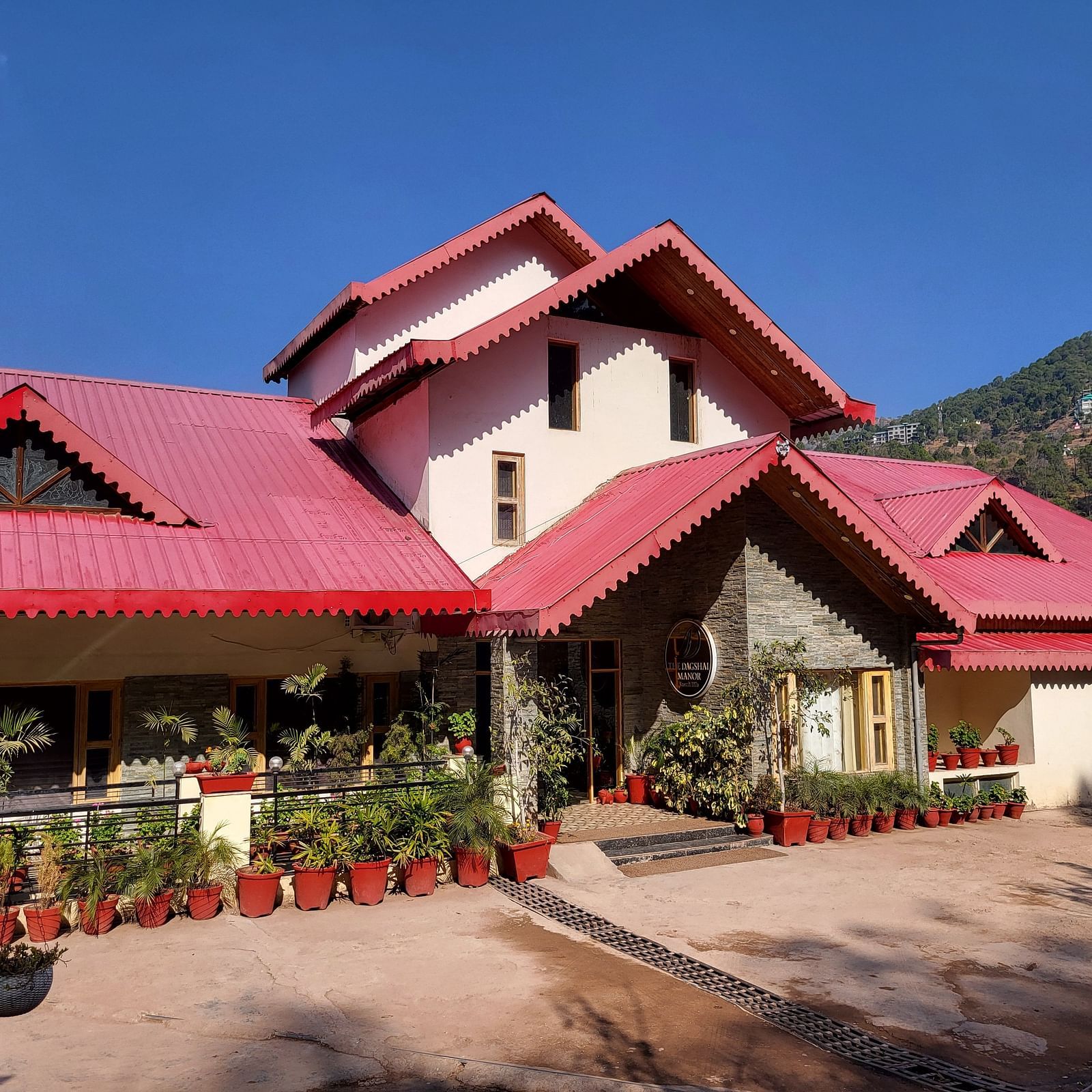 Facade of Dagshai Manor Kasauli - Inde Hotels with red roofs and potted plants under a clear blue sky.