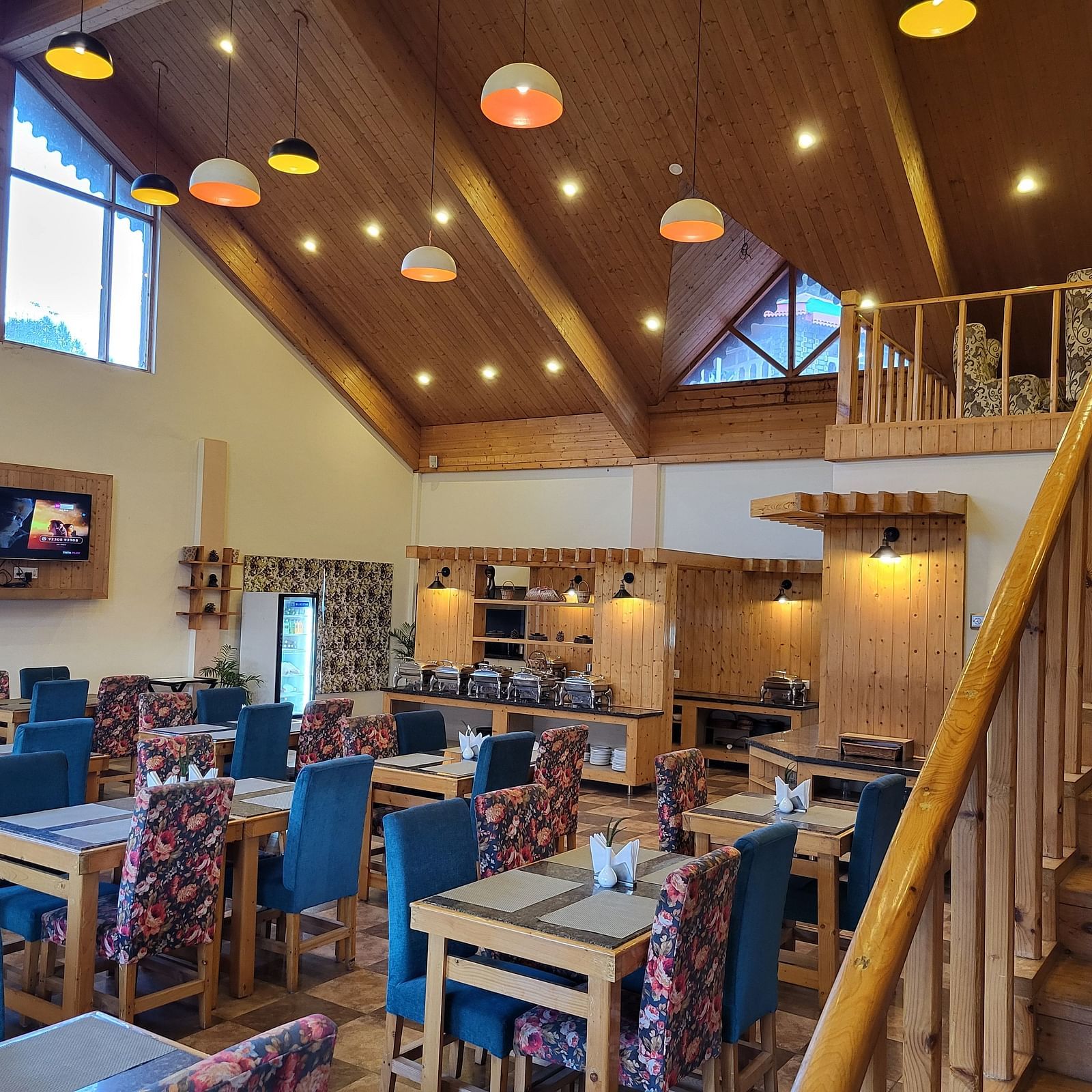 A well-lit, high-ceiling restaurant with wooden interiors, blue cushioned chairs, and an open staircase at Dagshai Manor Kasauli - Inde Hotels.