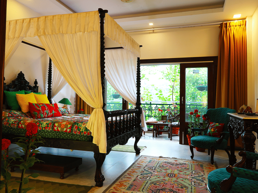 The Parakeet Room at Shaheen Bagh featuring a bed with canopy, wooden tables and chairs with plush cushions, and a balcony facing the lush greenery of the property.