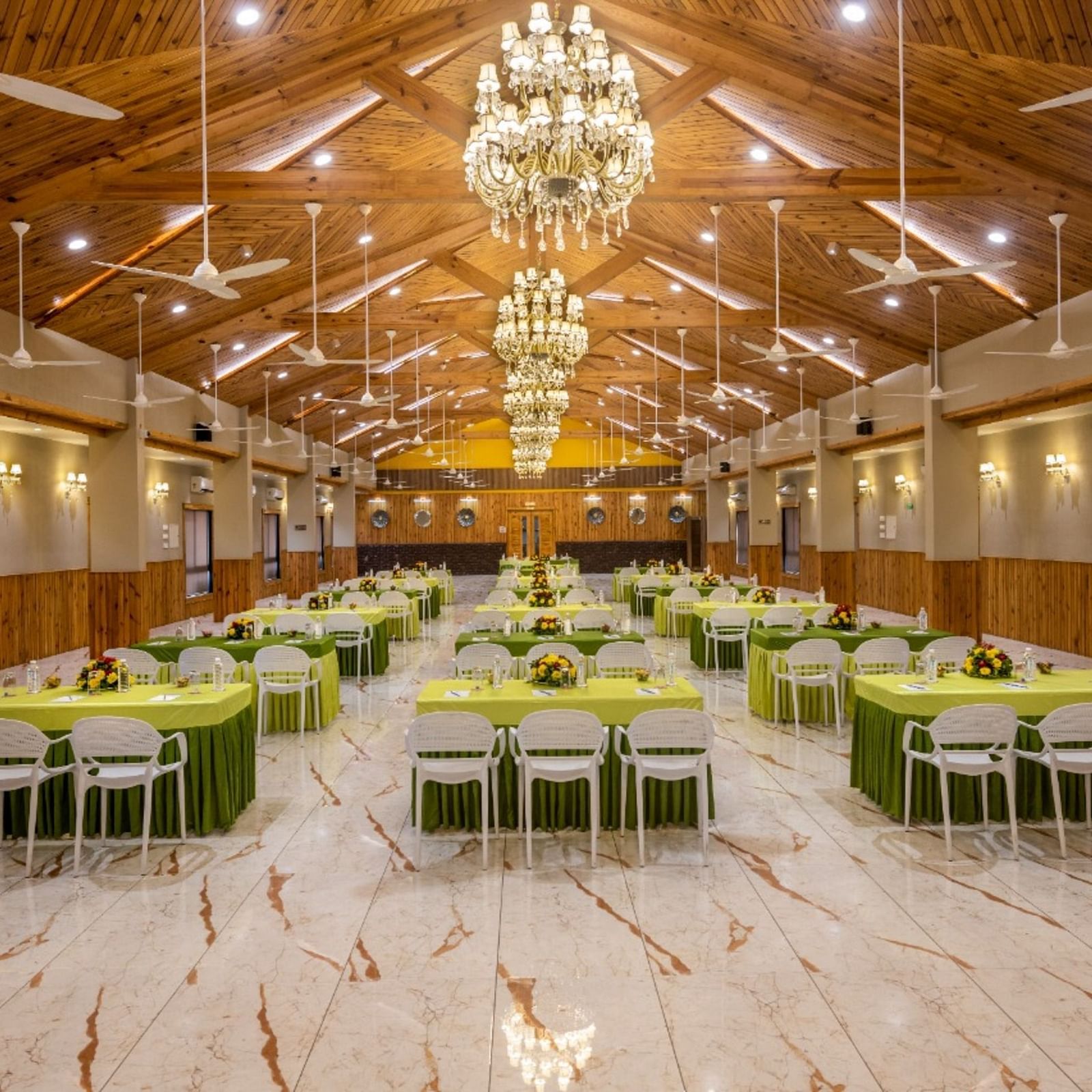 A well-lit banquet hall with multiple tables and chairs set for an event at Angel Lords Eco Inn Amreli.