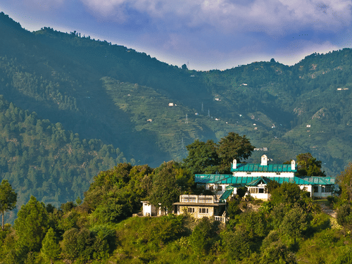 Facade of The Ramgarh Bungalows perched on a hill surrounded by scenic mountain view captured during the day.