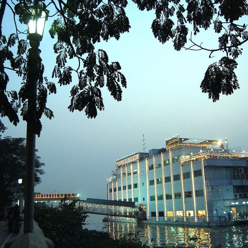 An exterior shot of a floating building with blue lighting, taken at dusk, showing a calm water body and trees in the foreground.
