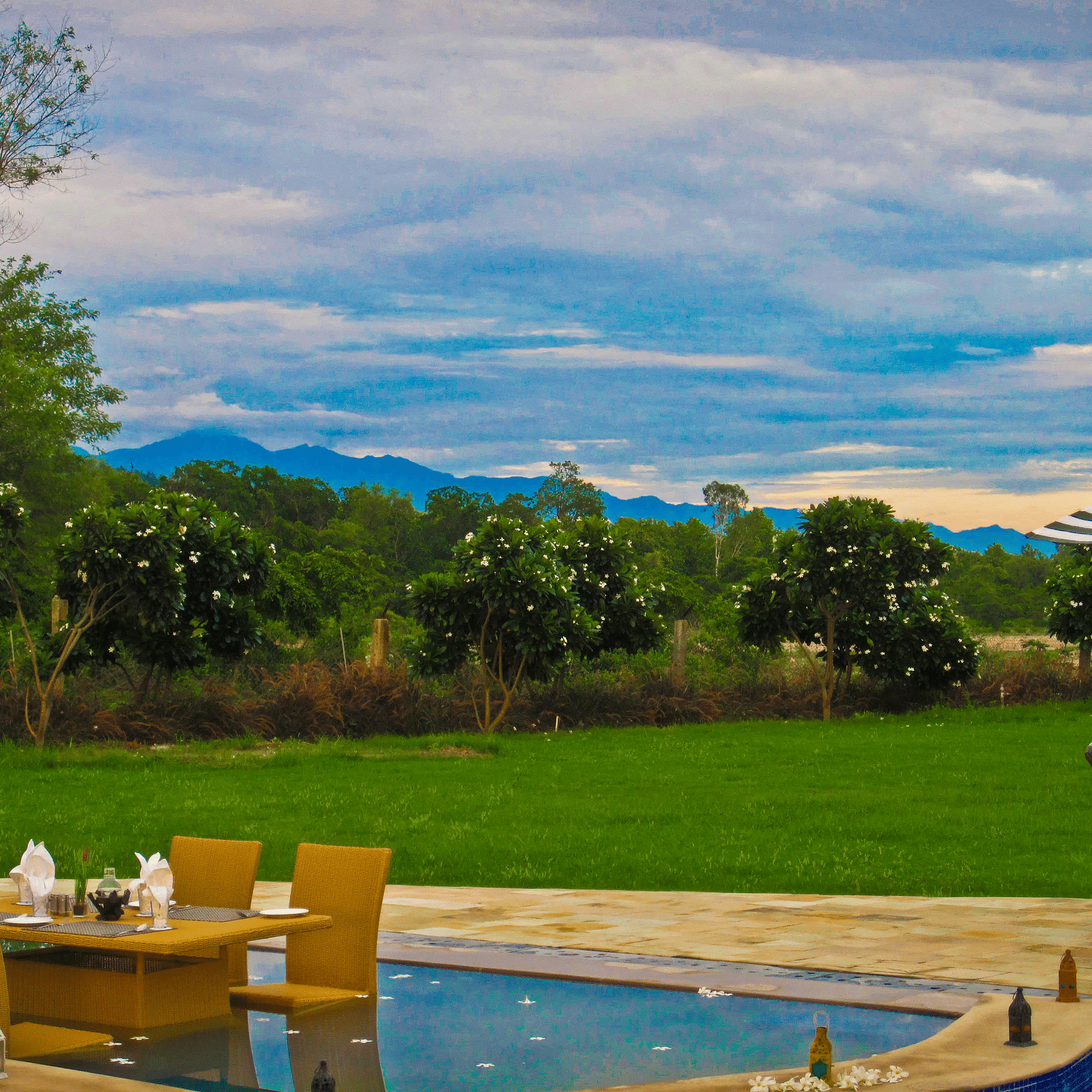 Full view shot of a lush green expansive lawn with a dining area half submerged in the swimming pool in the foreground - Ivory Destinations