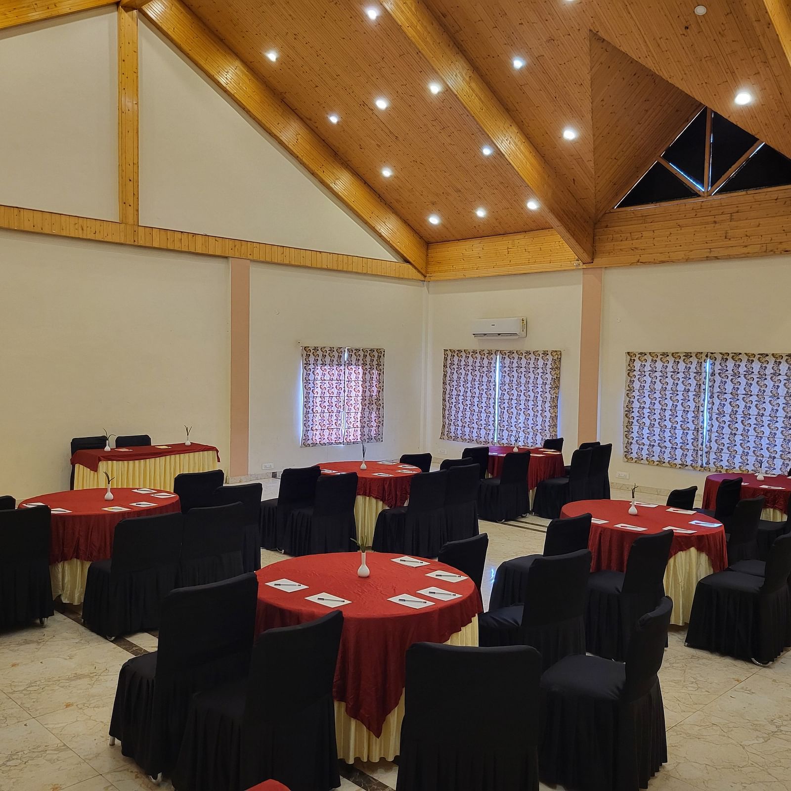 A spacious banquet hall with wooden ceiling beams, round tables, and elegant black and red seating at Dagshai Manor Kasauli - Inde Hotels.