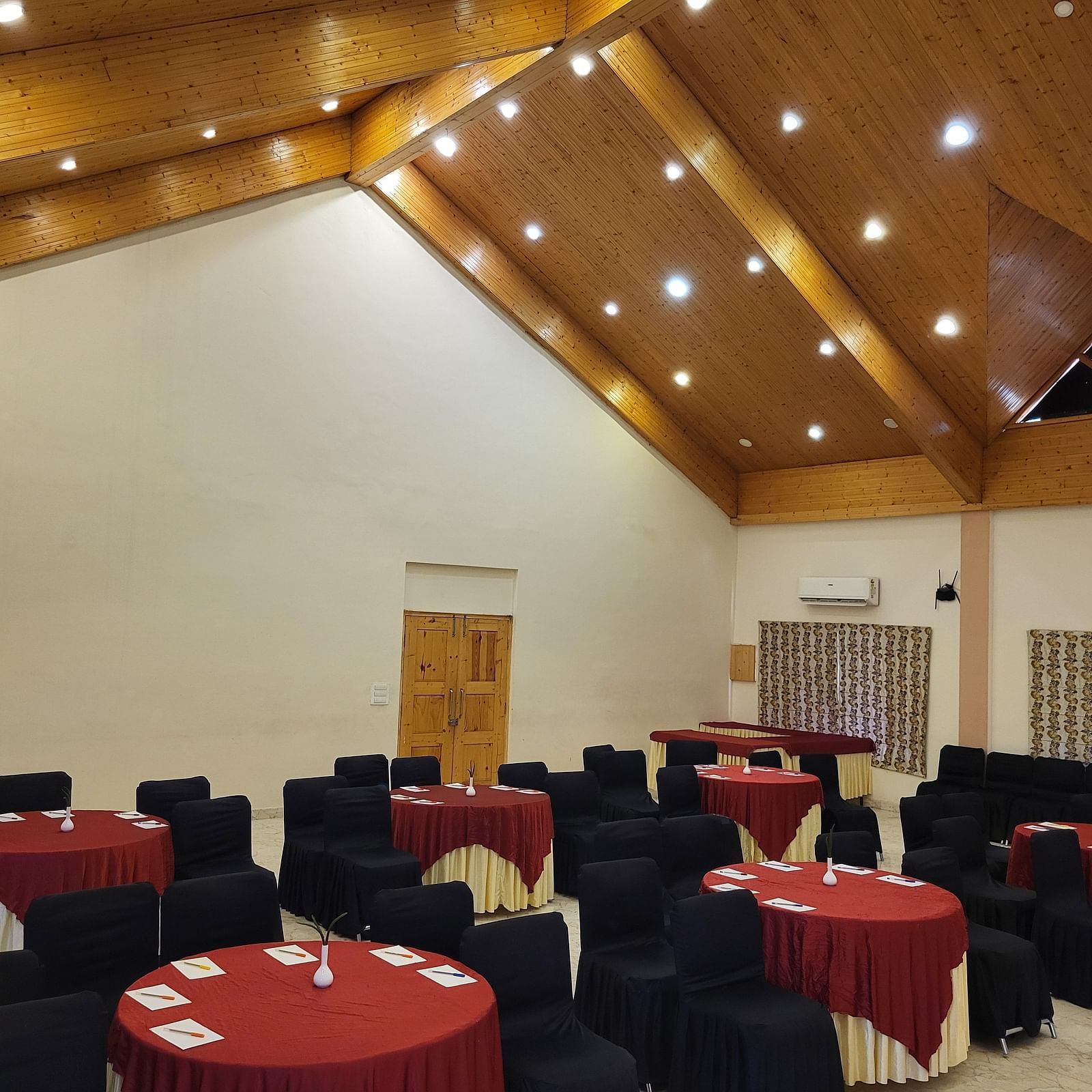 A spacious banquet hall with high wooden ceilings, elegant lighting, and round tables set for events at Dagshai Manor Kasauli - Inde Hotels.