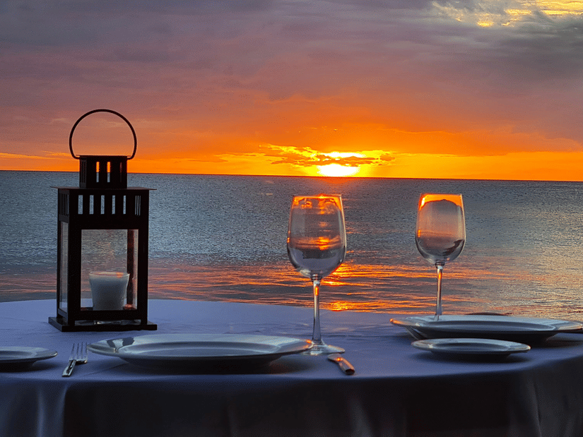 Romantic private dinner table for two with wine glasses and a lantern on the beach during a vibrant orange sunset