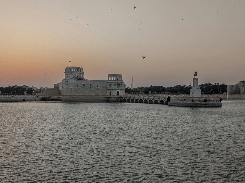 A view of a white building connected by a walkway over a lake, with another small white structure and surrounding buildings visible across the water.